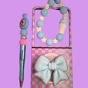 Pu&ograve; includere: Una penna metallica con un topper di perline che presenta un ciondolo a forma di personaggio blu, perline rosa e blu e un distanziatore trasparente. Accanto, una custodia rettangolare con motivo a scacchi rosa e bianco, un grande fiocco azzurro e un cinturino di perline.