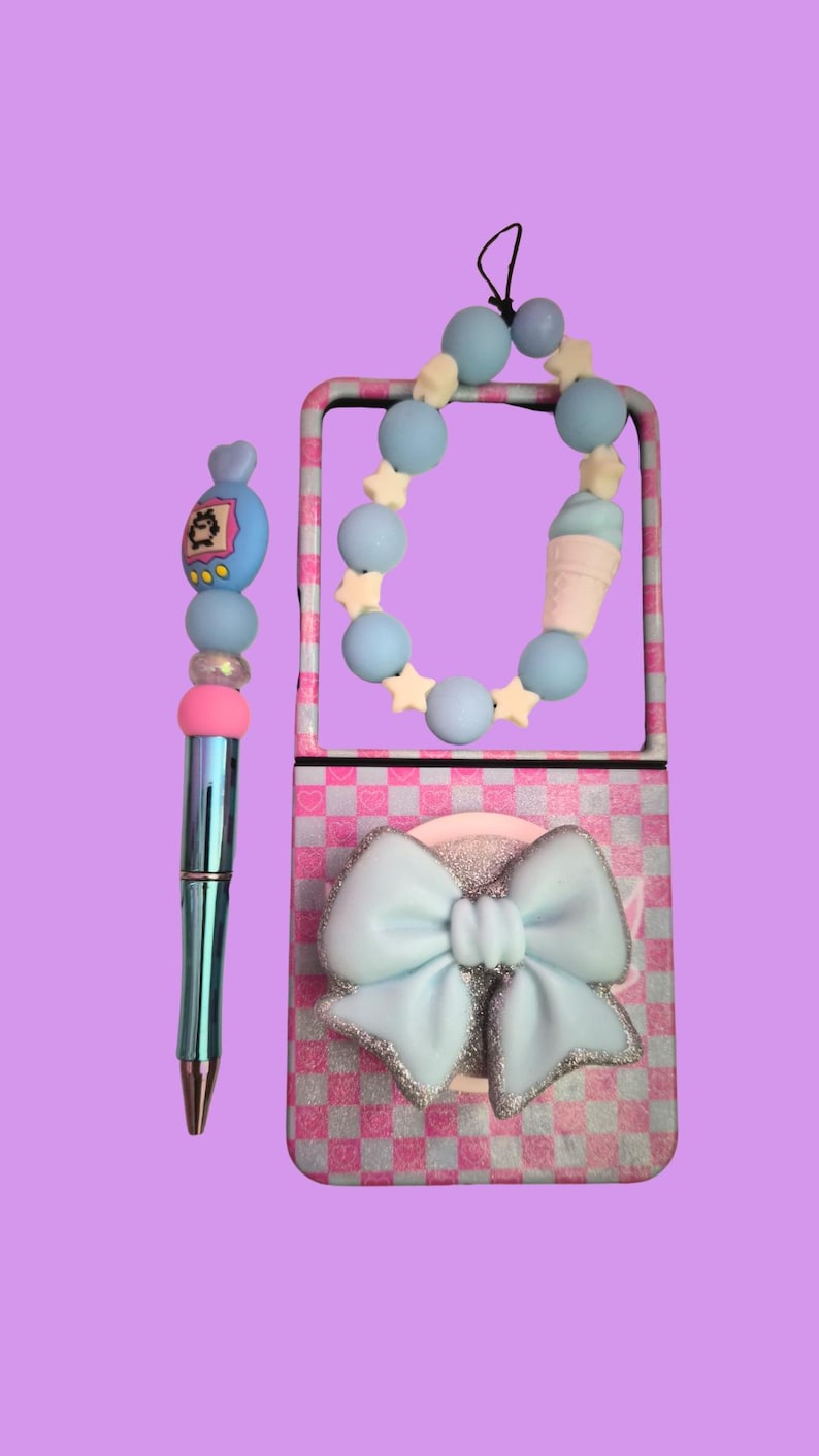 Pu&ograve; includere: Una penna metallica con un topper di perline che presenta un ciondolo a forma di personaggio blu, perline rosa e blu e un distanziatore trasparente. Accanto, una custodia rettangolare con motivo a scacchi rosa e bianco, un grande fiocco azzurro e un cinturino di perline.