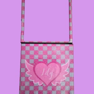 Pu&ograve; includere: Una custodia per telefono pieghevole con un motivo a scacchi rosa e bianco e una finitura glitterata. Il centro mostra un grande cuore rosa con ali d'angelo bianche e il nome Lily scritto in corsivo bianco sulla superficie del cuore.