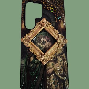 Pu&ograve; includere: Una cover per telefono rettangolare con uno sfondo scuro a motivi stellati e una cornice dorata decorata al centro. All'interno della cornice c'&egrave; un'illustrazione di due figure. La cover mostra anche un'immagine stampata pi&ugrave; grande e parzialmente visibile di due figure con abiti elaborati verdi e dorati.