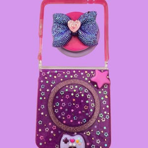 Pu&ograve; includere: Una custodia pieghevole per telefono viola decorata con piccole stelle colorate, un ciondolo a forma di stella rosa e una piccola figura di controller di gioco color lavanda. La parte superiore presenta un fiocco blu scuro glitterato.