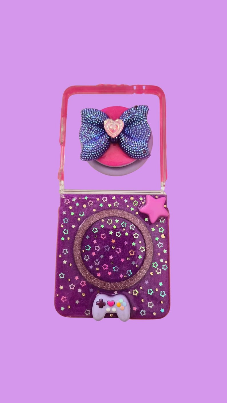 Pu&ograve; includere: Una custodia pieghevole per telefono viola decorata con piccole stelle colorate, un ciondolo a forma di stella rosa e una piccola figura di controller di gioco color lavanda. La parte superiore presenta un fiocco blu scuro glitterato.