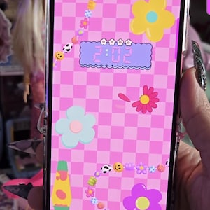 Può includere: Lo schermo di uno smartphone mostra un motivo a scacchi rosa con fiori dei cartoni animati, faccine sorridenti e una lampada a lava. L'ora 2:02 è visualizzata in un orologio digitale. Il telefono è in una custodia trasparente.