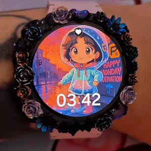 May include: A round smartwatch with a light pink strap and a black bezel decorated with small black and iridescent floral accents. The digital display shows a cartoon character in a raincoat with the text Happy Monday Motivation and 03:42.