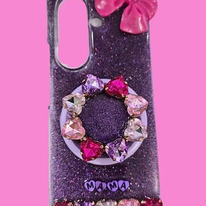 Pu&ograve; includere: Una cover per telefono viola glitterata con un fiocco rosa in alto, un supporto circolare decorato con cristalli a forma di cuore nelle tonalit&agrave; del rosa e del viola, la scritta MAMA in lettere viola e una fila di cinque cristalli in basso.