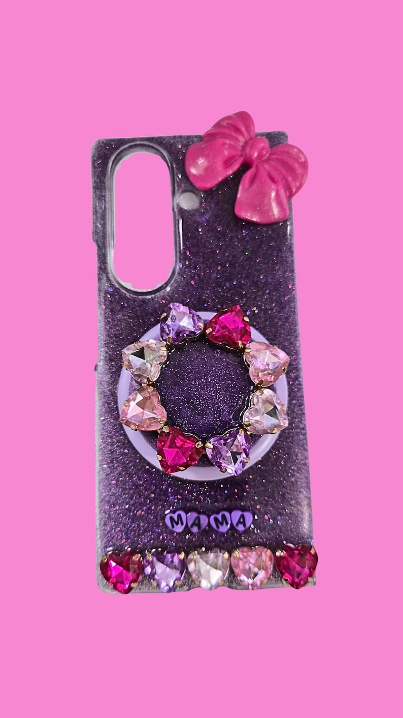 Pu&ograve; includere: Una cover per telefono viola glitterata con un fiocco rosa in alto, un supporto circolare decorato con cristalli a forma di cuore nelle tonalit&agrave; del rosa e del viola, la scritta MAMA in lettere viola e una fila di cinque cristalli in basso.