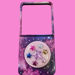 Pu&ograve; includere: Uno specchietto da borsa rettangolare con una stampa galassia blu e viola scuro con piccole stelle bianche. La parte inferiore presenta un inserto circolare in resina trasparente contenente diverse piccole decorazioni a forma di stella.