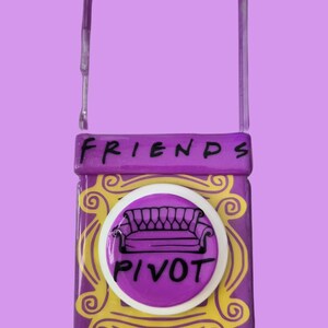 Pu&ograve; includere: Una cover per telefono rettangolare viola con un design a cornice decorativa gialla. La parola FRIENDS &egrave; stampata in nero nella parte superiore. &Egrave; presente un supporto circolare bianco, che mostra un disegno a linee nere di un divano e la parola PIVOT.
