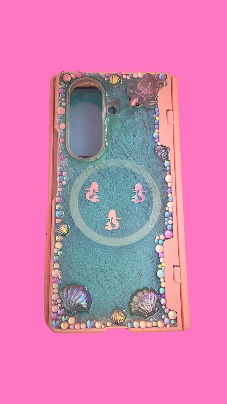 Pu&ograve; includere: Una cover per telefono rosa con un pannello centrale testurizzato color verde acqua e tre piccole sagome di sirene rosa. Il bordo &egrave; decorato con strass iridescenti, vari ciondoli a forma di conchiglia e una piccola tartaruga.