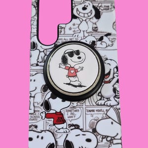 Pu&ograve; includere: Una custodia rettangolare per telefono con un motivo a fumetti in bianco e nero dei personaggi di Snoopy. Un supporto circolare nero per telefono &egrave; fissato al centro, mostrando un grafico di Snoopy con occhiali da sole e maglietta rossa.