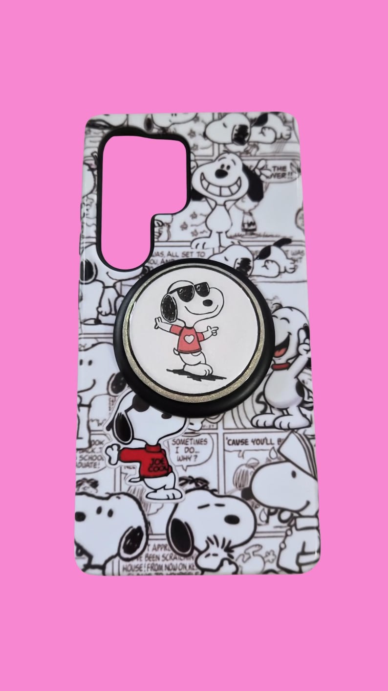 Pu&ograve; includere: Una custodia rettangolare per telefono con un motivo a fumetti in bianco e nero dei personaggi di Snoopy. Un supporto circolare nero per telefono &egrave; fissato al centro, mostrando un grafico di Snoopy con occhiali da sole e maglietta rossa.