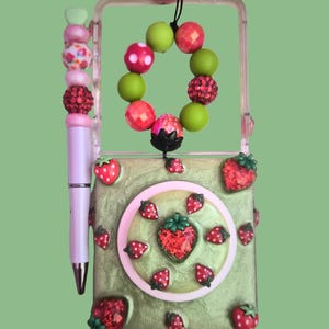 Pu&ograve; includere: Una custodia rettangolare verde chiaro decorata con piccoli ciondoli a forma di fragola rossa e un accento circolare centrale. Una penna rosa chiaro con un topper di perline &egrave; fissata sul lato. Un anello di perline con sfere verdi e rosa pende dall'alto.