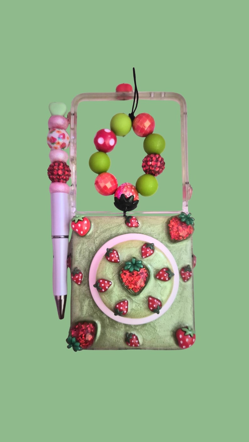 Pu&ograve; includere: Una custodia rettangolare verde chiaro decorata con piccoli ciondoli a forma di fragola rossa e un accento circolare centrale. Una penna rosa chiaro con un topper di perline &egrave; fissata sul lato. Un anello di perline con sfere verdi e rosa pende dall'alto.
