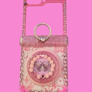 Pu&ograve; includere: Una custodia per telefono rosa con finitura glitterata, strass incastonati lungo i bordi e un'impugnatura circolare centrale. L'impugnatura &egrave; decorata con un'illustrazione di falena circondata da piccole rose rosa e un anello in metallo.