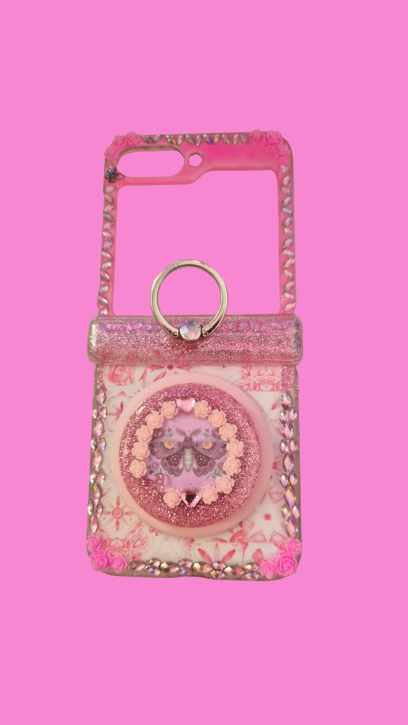 Pu&ograve; includere: Una custodia per telefono rosa con finitura glitterata, strass incastonati lungo i bordi e un'impugnatura circolare centrale. L'impugnatura &egrave; decorata con un'illustrazione di falena circondata da piccole rose rosa e un anello in metallo.