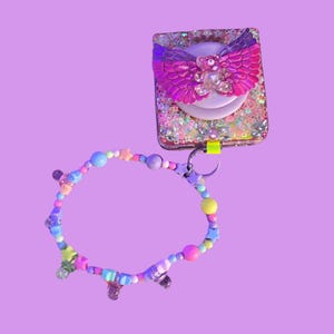 Può includere: Un supporto per telefono con un design ad ali di farfalla rosa e un braccialetto di perline con perline colorate e ciondoli a forma di stella. Il supporto per telefono è decorato con glitter e una piccola figura di orso. Il braccialetto ha una varietà di colori pastello.