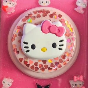 May include: A pink square compact mirror featuring a central circular design with a white Hello Kitty face and a pink bow. The surrounding area is filled with iridescent heart-shaped sequins, and the corners feature small Hello Kitty and Kuromi figures.
