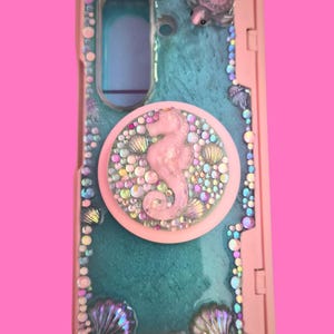 Pu&ograve; includere: Una cover per telefono rettangolare con superficie in resina color ottanio, decorata con strass iridescenti e ciondoli a forma di conchiglia. &Egrave; presente un supporto circolare rosa con un cavalluccio marino centrale e piccoli cristalli.