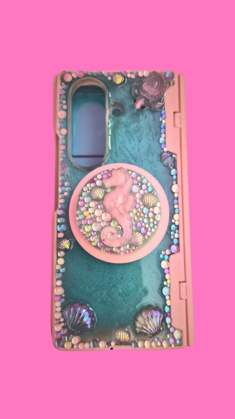 Pu&ograve; includere: Una cover per telefono rettangolare con superficie in resina color ottanio, decorata con strass iridescenti e ciondoli a forma di conchiglia. &Egrave; presente un supporto circolare rosa con un cavalluccio marino centrale e piccoli cristalli.