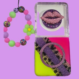 Pu&ograve; includere: Una cover quadrata per telefono con base rosa acceso e giallo neon, caratterizzata da una striscia diagonale glitterata viola, una fascia con stampa leopardata nera e oro e un anello. Un decoro a labbra in strass rosa 3D e un cinturino di perline.