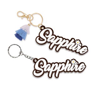 May include: Two keychains with the name "Sapphire" in a white, cursive font with a brown outline. One keychain has a gold clasp and a blue tassel. The other keychain has a silver chain and ring.