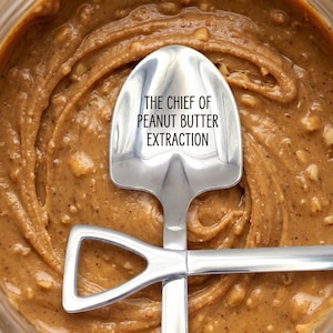 May include: A close-up shot of peanut butter in a jar with a stainless steel spoon. The spoon has the text "THE CHIEF OF PEANUT BUTTER EXTRACTION" engraved on it. The peanut butter has a creamy, textured appearance.