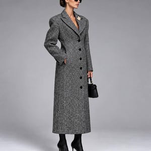 May include: A long, grey and black herringbone patterned wool coat featuring a notched lapel, structured shoulders, and a single-breasted front with six black buttons. The coat includes a floral brooch on the lapel, side pockets, and long sleeves.