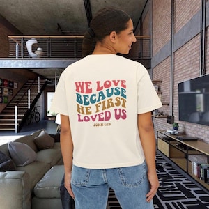 Könnte beinhalten: Ein cremefarbenes Kurzarm-T-Shirt mit einer mehrfarbigen, wellenförmigen Grafik auf der Rückseite. Der Text WE LOVE BECAUSE HE FIRST LOVED US JOHN 4:19 ist in gestapelten Buchstaben im Retro-Stil in Rosa, Petrol und Senfgelb aufgedruckt.