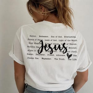 Könnte beinhalten: Ein weißes T-Shirt mit schwarzem Text auf der Rückseite. Das Wort Jesus ist in einer großen Schreibschrift in der Mitte geschrieben, umgeben von kleineren gedruckten Wörtern wie Savior, Redeemer, Son of God, Everlasting, Prince of Peace und einem Herz.