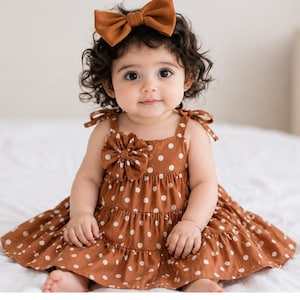 May include: A baby wearing a brown polka dot dress with a matching bow headband. The dress has a tiered skirt and a large bow detail on the bodice. The baby is sitting on a white surface.