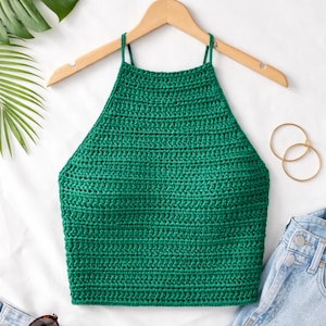 May include: A green crochet halter top with thin straps hangs on a wooden hanger. The top is made of a textured, knitted material. Gold hoop earrings and denim jeans are visible in the background.