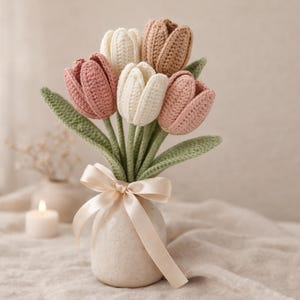 Op de afbeelding: Een boeket gehaakte tulpen in een kleine, getextureerde vaas. De bloemen zijn in tinten roze, crème en beige, met groene bladeren. Een crèmekleurig lint is om de vaas gebonden. De achtergrond is een zachte, neutrale kleur.