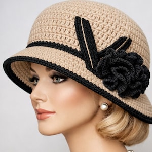May include: A beige crochet cloche hat with a black brim and a black floral accent. The hat has a rounded crown and a small brim, with a black decorative band around the base of the crown.