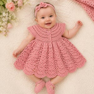 Pode incluir: Um bebé a usar um vestido de croché rosa claro feito à mão, uma faixa a condizer e sapatos. O vestido apresenta uma bainha com vieiras e fecho de botão. O bebé está deitado numa superfície branca e macia, com flores cor-de-rosa no fundo.
