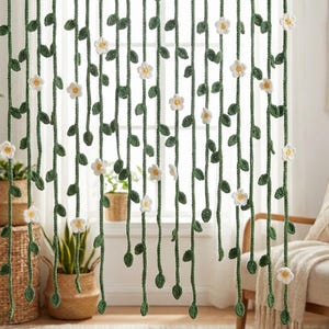 May include: Crocheted curtain with a floral design. The curtain features vertical green vines with attached green leaves and white flowers. The curtain hangs in front of a window, with plants in baskets visible in the background.
