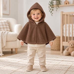 Op de afbeelding: Een bruine fleece cape met capuchon en ritssluiting aan de voorzijde, gedragen over een crèmekleurig shirt met lange mouwen en een lichtbeige broek, gecombineerd met bijpassende beige schoenen. Het kledingstuk heeft een zachte textuur.