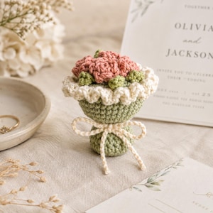 May include: A small, handcrafted crochet bouquet featuring dusty pink roses and green leaves, wrapped in a sage green base with a cream-colored ruffled edge and a tied cream yarn bow. The item is a decorative piece with a textured, yarn-based construction.