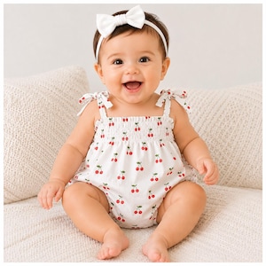 Peut inclure: Un bébé porte un barboteuse blanc orné d'un motif de cerises rouges et un bandeau blanc avec un nœud. La barboteuse a des bretelles nouées et un corsage smocké. Le bébé sourit.