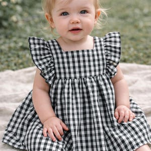 May include: A baby in a black and white gingham dress with frilled sleeves. The dress features a square neckline and a gathered waist. The baby is seated on a light-coloured blanket outdoors.