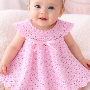 May include: A light pink crochet baby dress with a scalloped neckline and cap sleeves. A pink satin ribbon is tied in a bow at the waist. The dress is made of a soft, open-weave yarn.