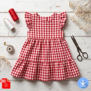 May include: A red and white gingham dress with frilled sleeves and a tiered skirt. The dress is on a white wooden surface, with sewing supplies including thread spools, a measuring tape, and scissors.