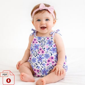 May include: A baby wearing a floral print romper with a pink bow headband. The romper features a white background with purple and pink flowers. The baby is smiling and sitting on a white surface.