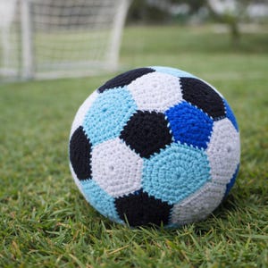 May include: A handmade crochet soccer ball with a classic design. The ball features a pattern of black, white, and various shades of blue hexagons. It rests on a bed of green grass, with a soccer goal in the background.