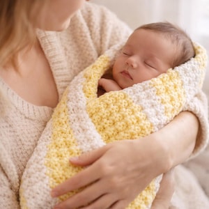 Op de afbeelding: Een slapende baby gewikkeld in een handgemaakte gehaakte deken. De deken heeft afwisselende strepen in geel en wit. De baby wordt in iemands armen gehouden en draagt een lichtgeleurde trui.