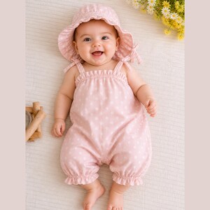 May include: A baby wearing a pink romper with white polka dots and a matching sun hat. The romper has spaghetti straps and ruffled hems. The hat has a wide brim and ties. The baby is smiling.