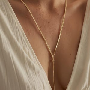 Puede incluir: Un collar dorado con un diseño de cadena delicado. El collar tiene un colgante en forma de Y que termina en un nudo. El collar se usa sobre una blusa blanca con escote.