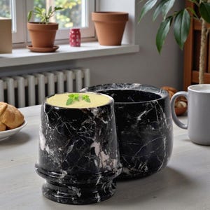 May include: Two black marble cups with white veining sit on a light-colored table. One cup is filled with a light green soup and garnished with a sprig of green herbs. A gray mug and a plate of bread are also on the table.