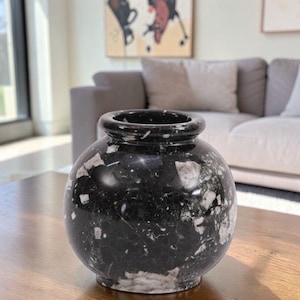 May include: A black marble vase with white and gray veining. The vase has a rounded body and a slightly flared rim. It sits on a wooden table in a well-lit room. The vase is a decorative item.