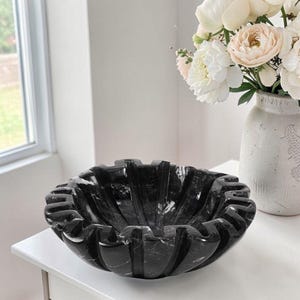 May include: A black marble decorative bowl with a scalloped edge. The bowl is sitting on a white surface. A vase of white and peach flowers is in the background.
