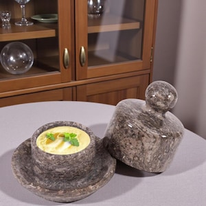 May include: A stone soup bowl with a saucer, filled with yellow soup and garnished with parsley. A matching stone lid sits beside the bowl. The bowl and lid are made of a speckled gray stone.